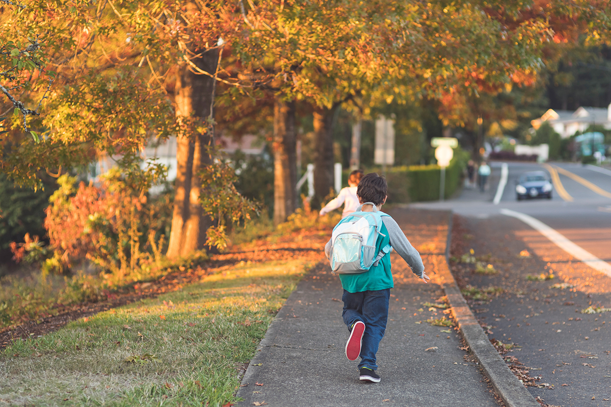 A young boy running home from school next to a road. 
