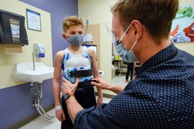 A child wearing a surgical mask stands as an adult wearing a surgical mask helps fit them for a scoliosis brace.