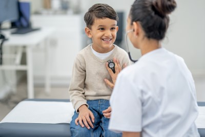  A young boy sits up on an exam table in a doctors office during a routine check-up. His doctor is seated in front of him and listening to his heart with her stethoscope.