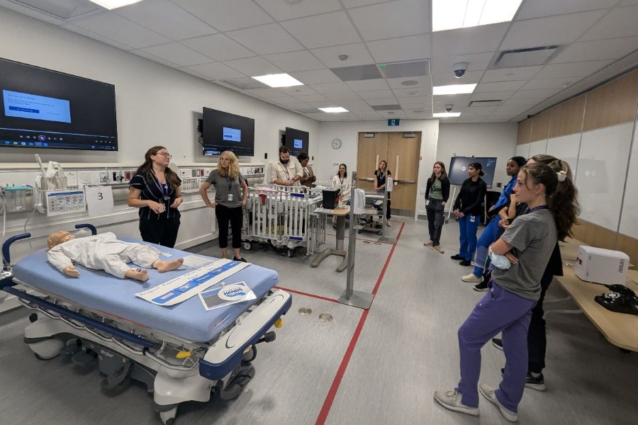 People in a medical training room with hospital beds, mannequins, and equipment, participating in a simulation session.