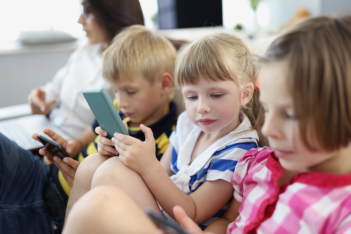 Three kids sit on a couch looking at phones, an adult is on a laptop.