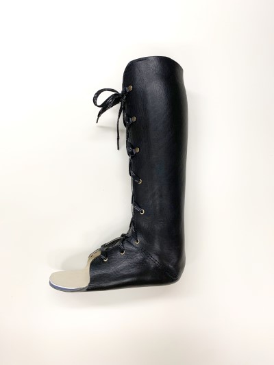 A tall leather black boot with laces up the front used for ankle and foot bracing is displayed in front of a white background.