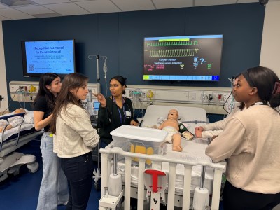 Four individuals participate in a clinical training session around a medical mannequin in a hospital-like environment.