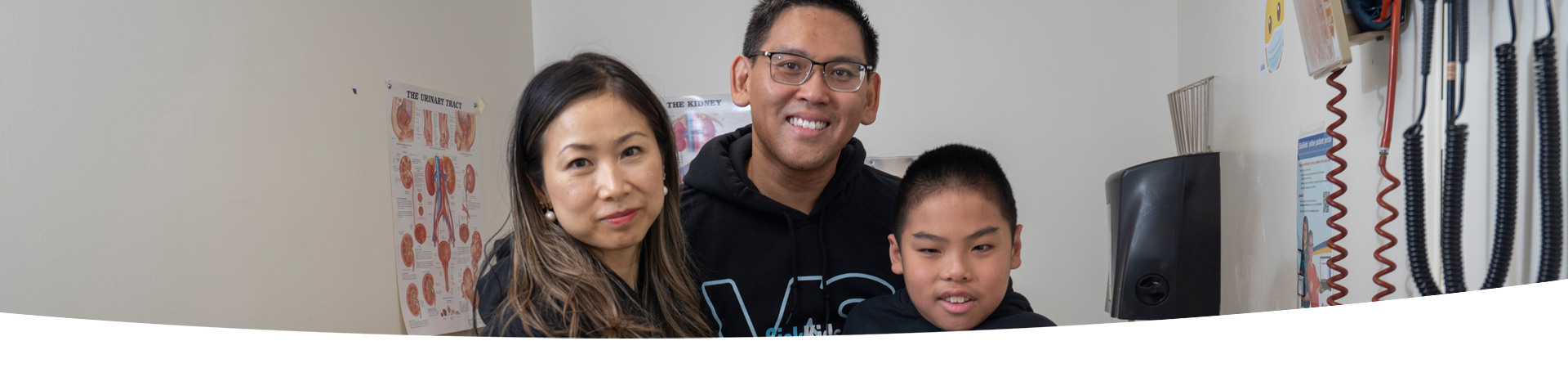 A family of three posing together for a photo at the Kidney Transplant Clinic. In the background, there are posters of kidney diagrams and medical equipment hanging from the walls.