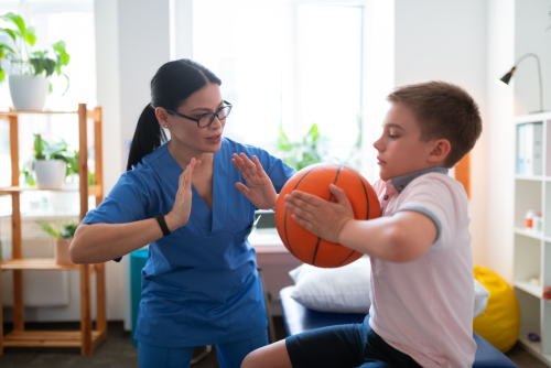 Young boy pushing his hands into a basketball as part of a physiotherapy exercise, instructed by a physiotherapist