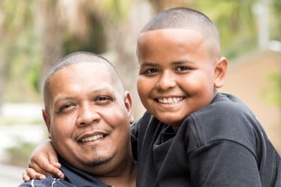 A father with his son perched on his shoulder smiling at the camera.