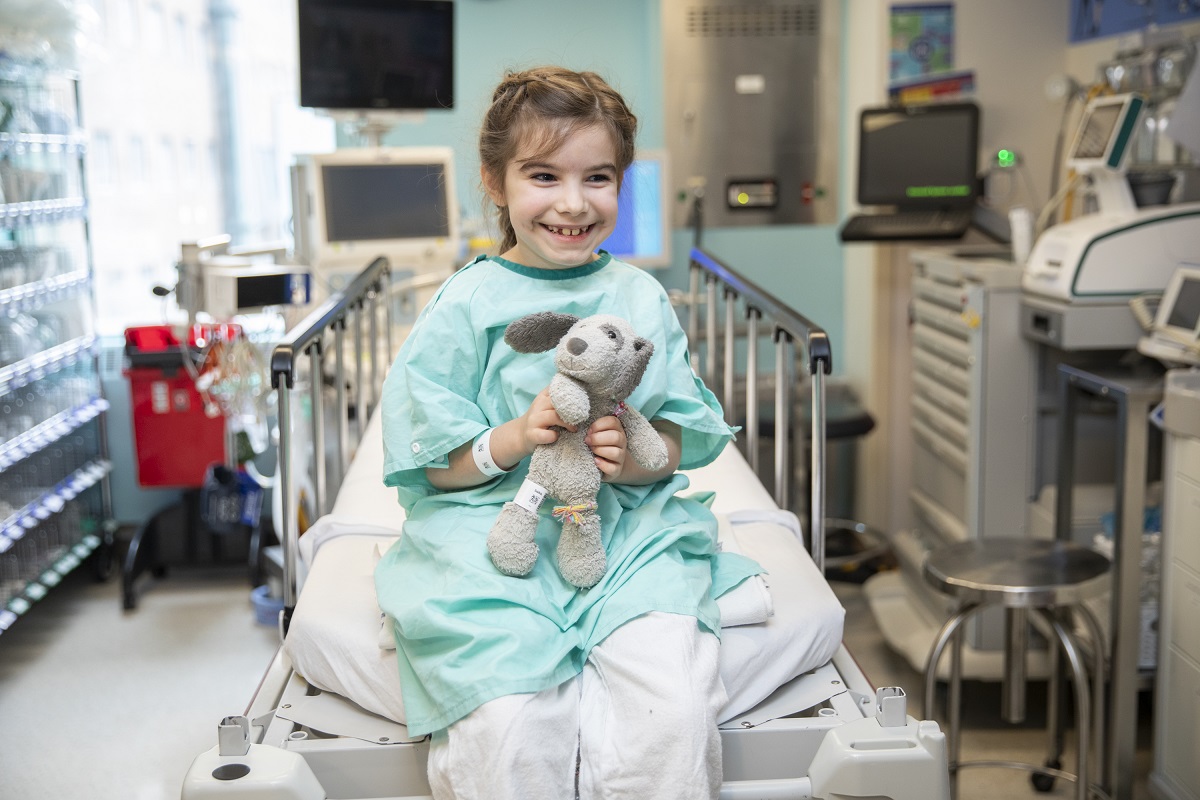 A child in a hospital gown sits on a bed holding a stuffed animal in a medical room.