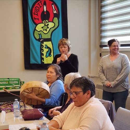 A group of individuals gathered in a well-lit room, participating in a meeting or workshop. One person stands while playing a large drum, contributing to a cultural or ceremonial moment.