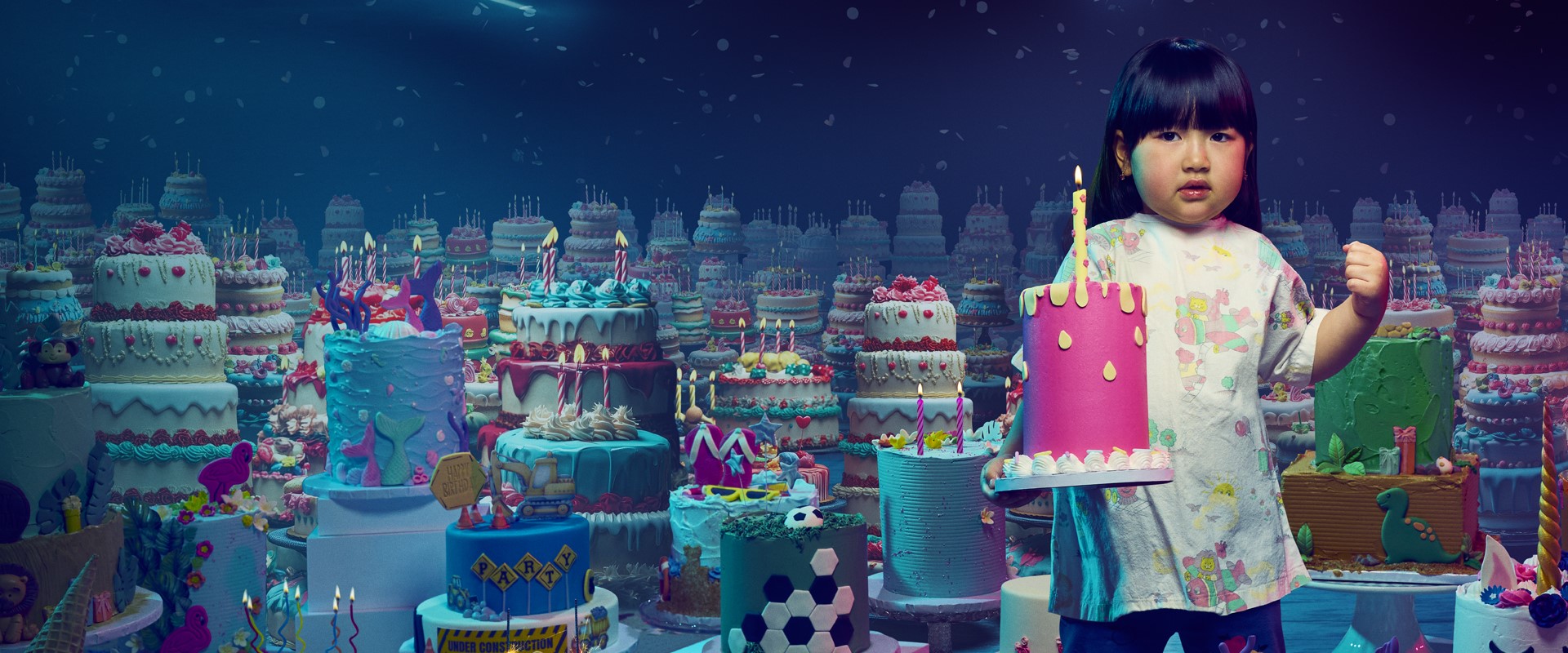 A girl stands holding a birthday cake, amongst a sea of birthday cakes.