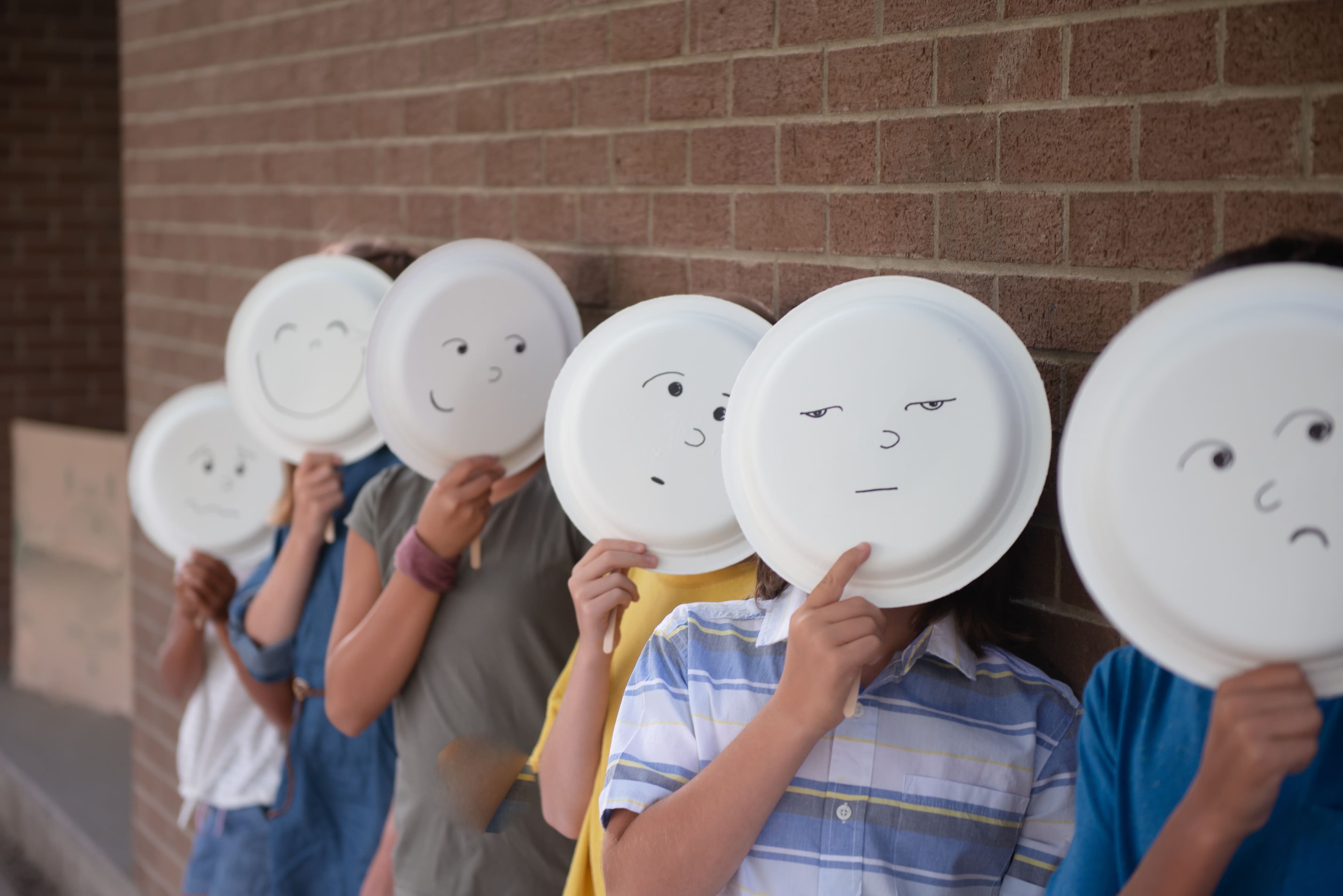 Children in a row holding paper plates over their faces. The paper plates have faces drawn on in marker.