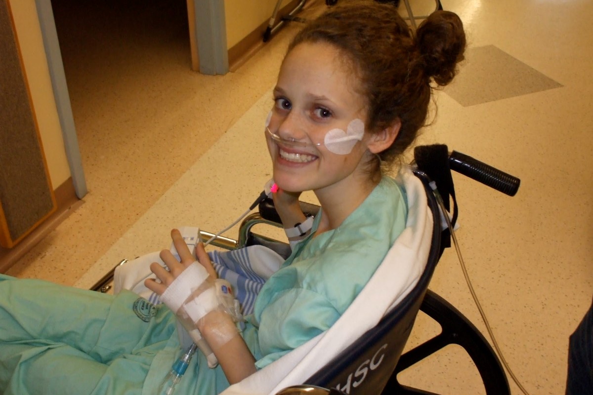 Young teenage girl with a bandage on her face smiling while seated in a wheelchair, wearing a hospital gown with medical tubes visible around her neck area.