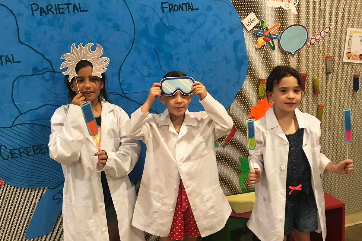Three children in white lab coats. They are holding paper crafts of test tubes and goggles. The wall behind them has science related decorations, such as a brain with its lobes labeled, beakers, DNA, microscopes and more.