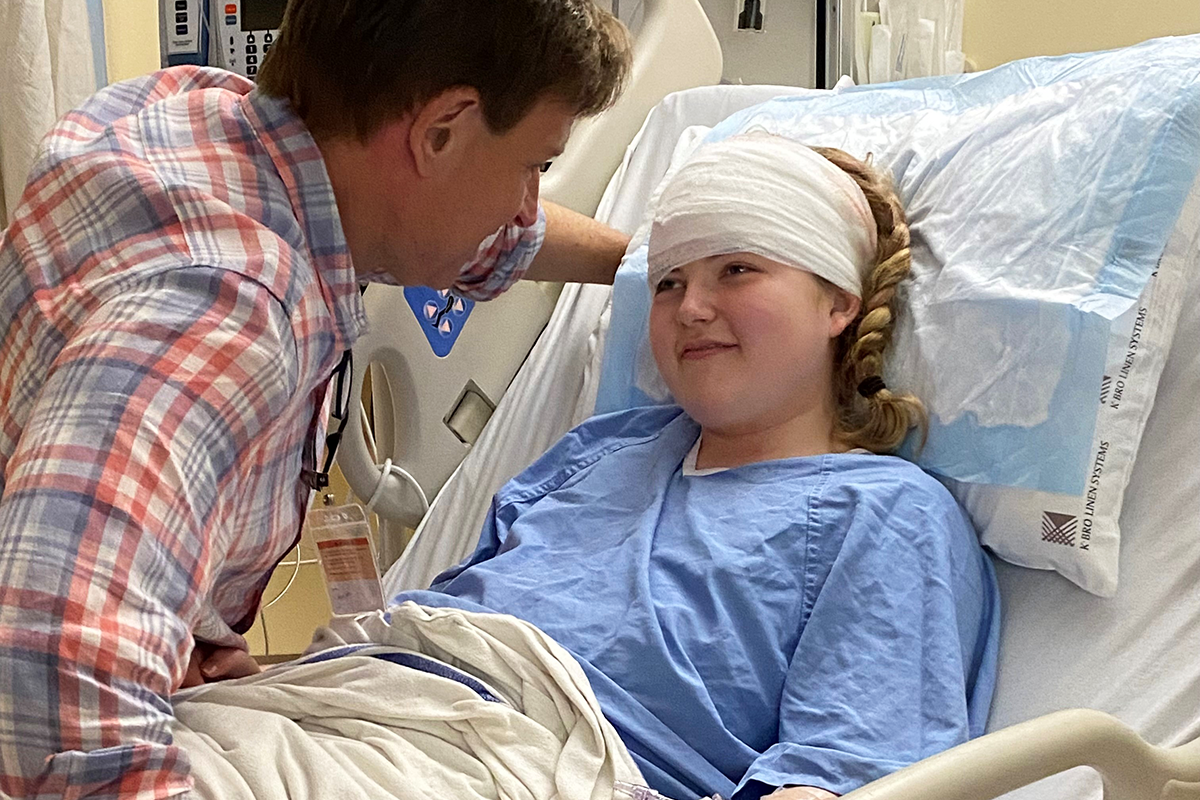 A young women lies in a hospital bed, her head bandaged after surgery, smiling at her father.