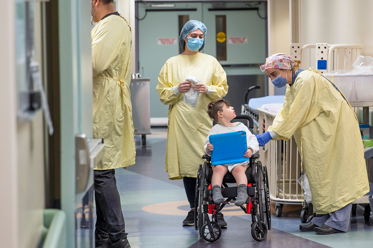 Child sits in a wheelchair holding a tablet. Three adults in medical gowns, caps and masks are present. One leans over and speaks to the child.