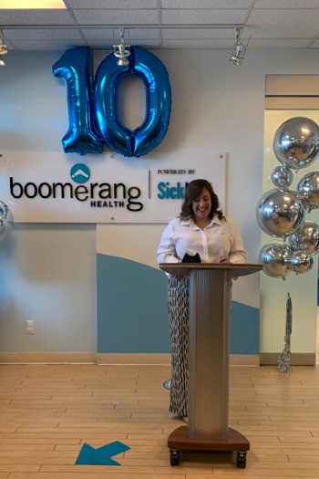 Alexandra Ieraci standing at a podium in front of a sign that says "Boomerang Health, powered by SickKids" and a set of blue and silver balloons, with the blue balloons in the shape of the number 10.