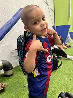 A young boy stands in a gym wearing a backpack with a tube attached to it.