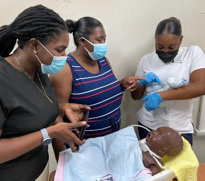 Medical training scene where three masked women observe a healthcare worker demonstrating infant care techniques using a baby mannequin.