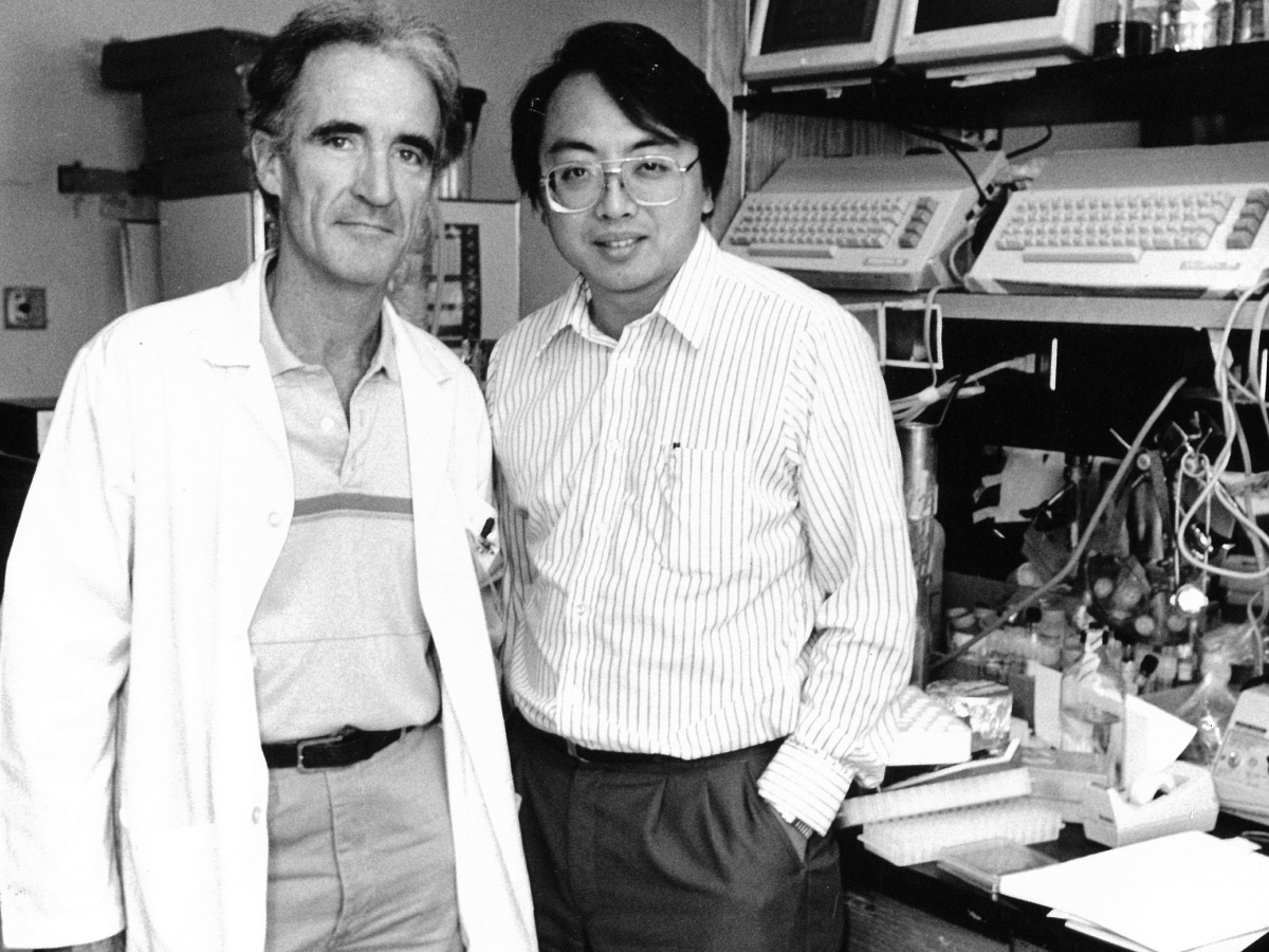 Two researchers in a medical lab - older man in lab coat and younger Asian colleague with computers behind them.