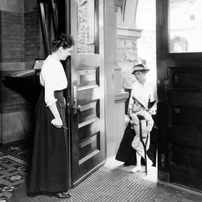 Historic black and white photo of woman greeting another woman with child in formal attire at doorway.