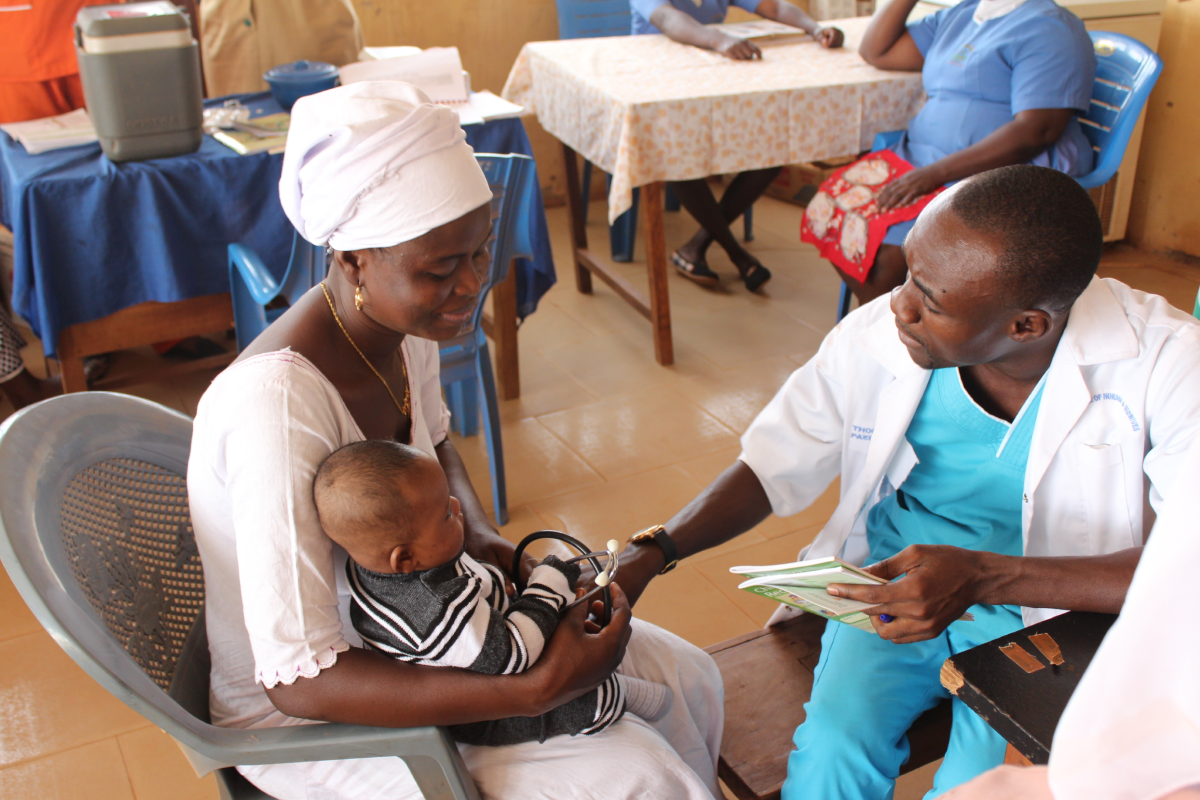 Healthcare worker in teal scrubs examining infant held by mother in white headwrap at rural clinic.