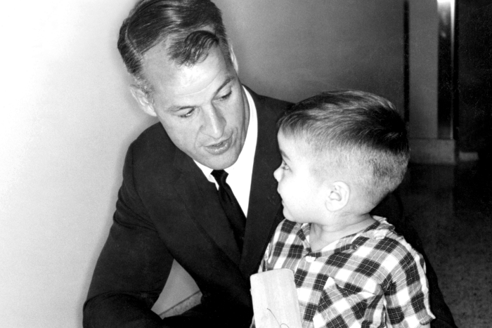 Black and white photo of man in suit kneeling beside young boy holding a crutch or wooden walking aid.