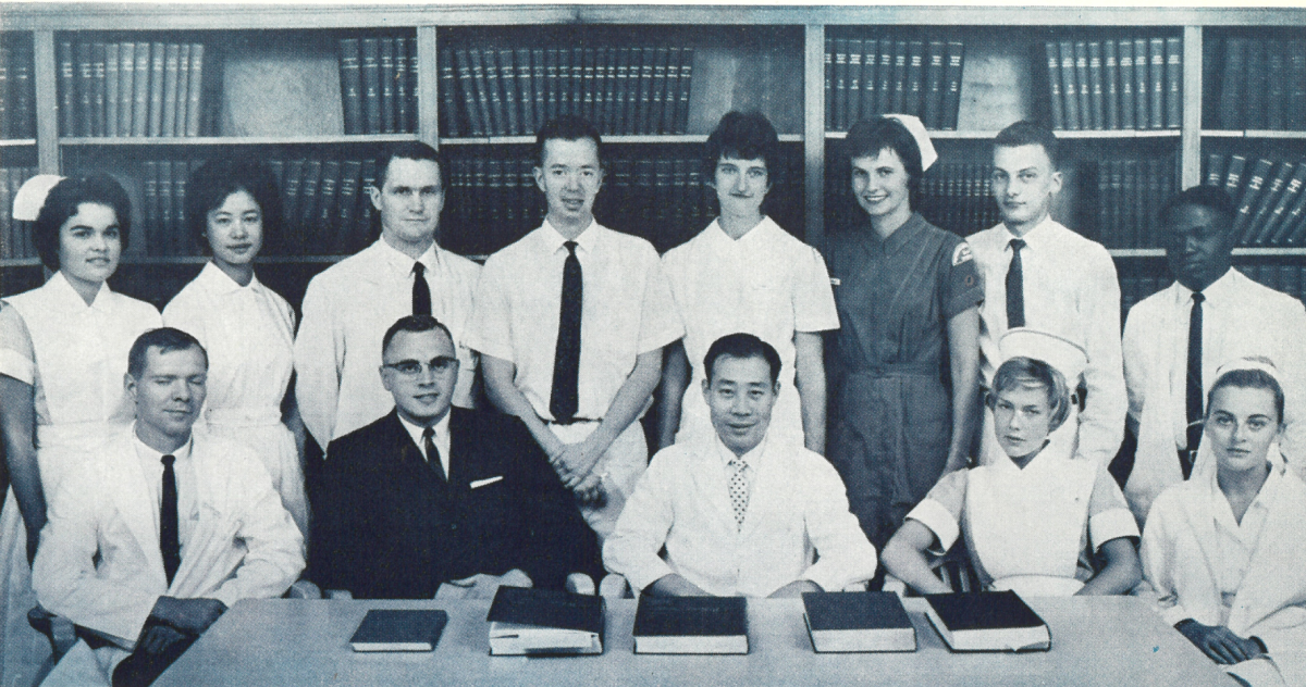 Black and white photograph captioned "H.S.C.'s Second Function is Education" showing a diverse group of medical students or staff. Includes nurses in uniforms with caps and medical personnel in white coats, arranged in two rows with bookshelves behind them and reference books on the table.