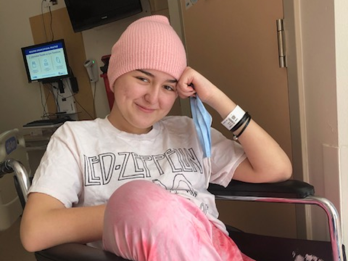  Young patient wearing a pink beanie hat and hospital wristband smiling in a hospital room. She's wearing a Led Zeppelin t-shirt and pink pants.