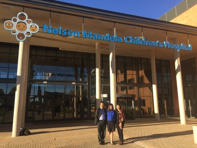 Three people stand at entrance of Nelson Mandela Children's Hospital with colorful children's faces logo above.