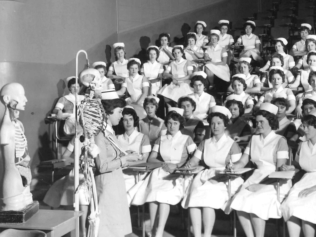 Black and white photograph of nursing students in a classroom setting with anatomical models. Rows of women in white uniforms and caps attend an anatomy lecture.