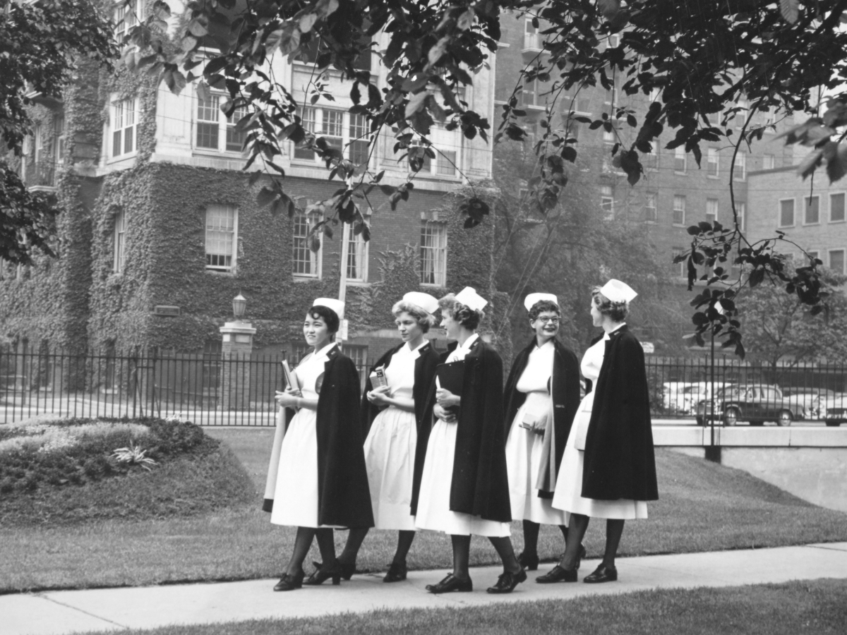 Five nurses in traditional uniforms with white caps and black capes walking together outside an ivy-covered hospital building in a black and white photograph.