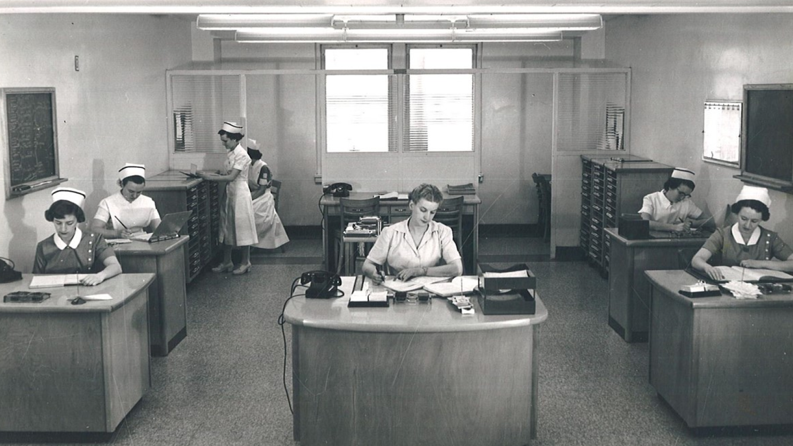 A vintage black and white photograph of a hospital nursing station with several nurses in white uniforms working at desks and filing cabinets.