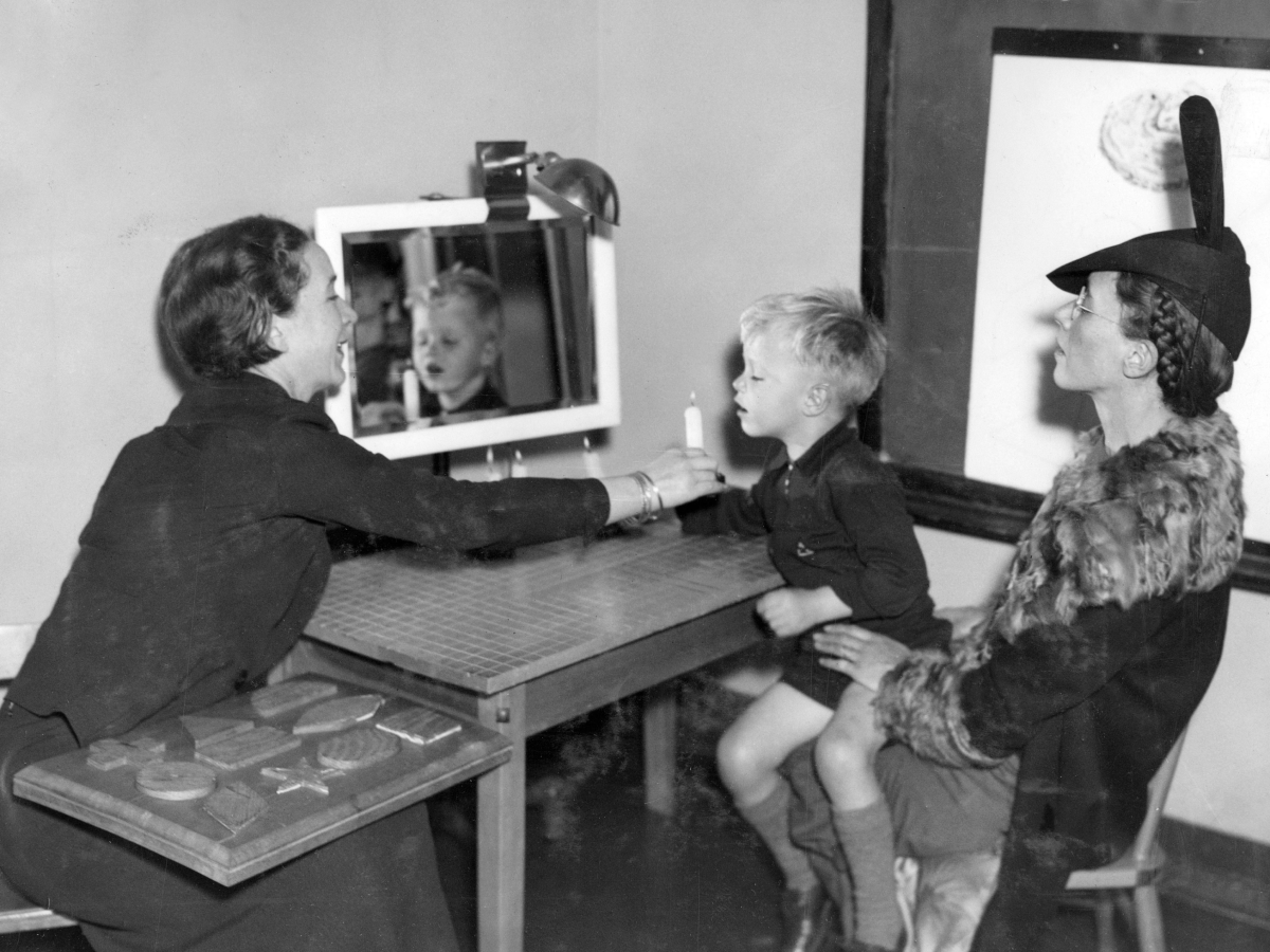 You're right - this is a speech therapy session. A professional is working with a young boy, using what appears to be some kind of audio-visual or speech therapy equipment, while a woman (likely his mother) in a fur coat and hat observes the session.