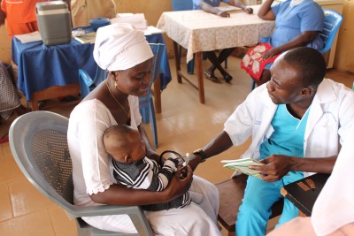 A healthcare worker examines an infant being held by its mother at what appears to be a rural health clinic or community health center. The mother wears a white head covering and dress, while the medical professional in a white coat and teal scrubs takes the baby's measurements. Other patients can be seen waiting at tables in the background.