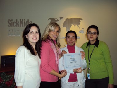 Four women pose at SickKids International Affairs office with a healthcare certificate recipient in white coat holding award.