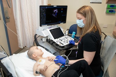 A child lies on a hospital bed with chest sensors while an adult uses an ultrasound probe to scan the chest. A heart image appears on a medical monitor.
