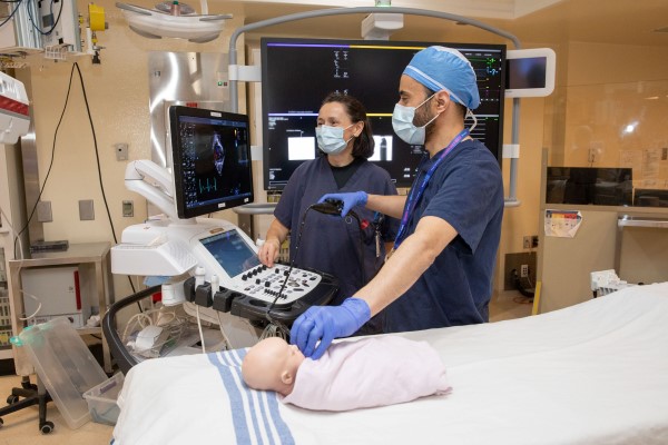 An infant mannequin on a hospital bed while two clinicians use an ultrasound probe to examine the chest. Heart images are shown on a medical monitor beside the bed.