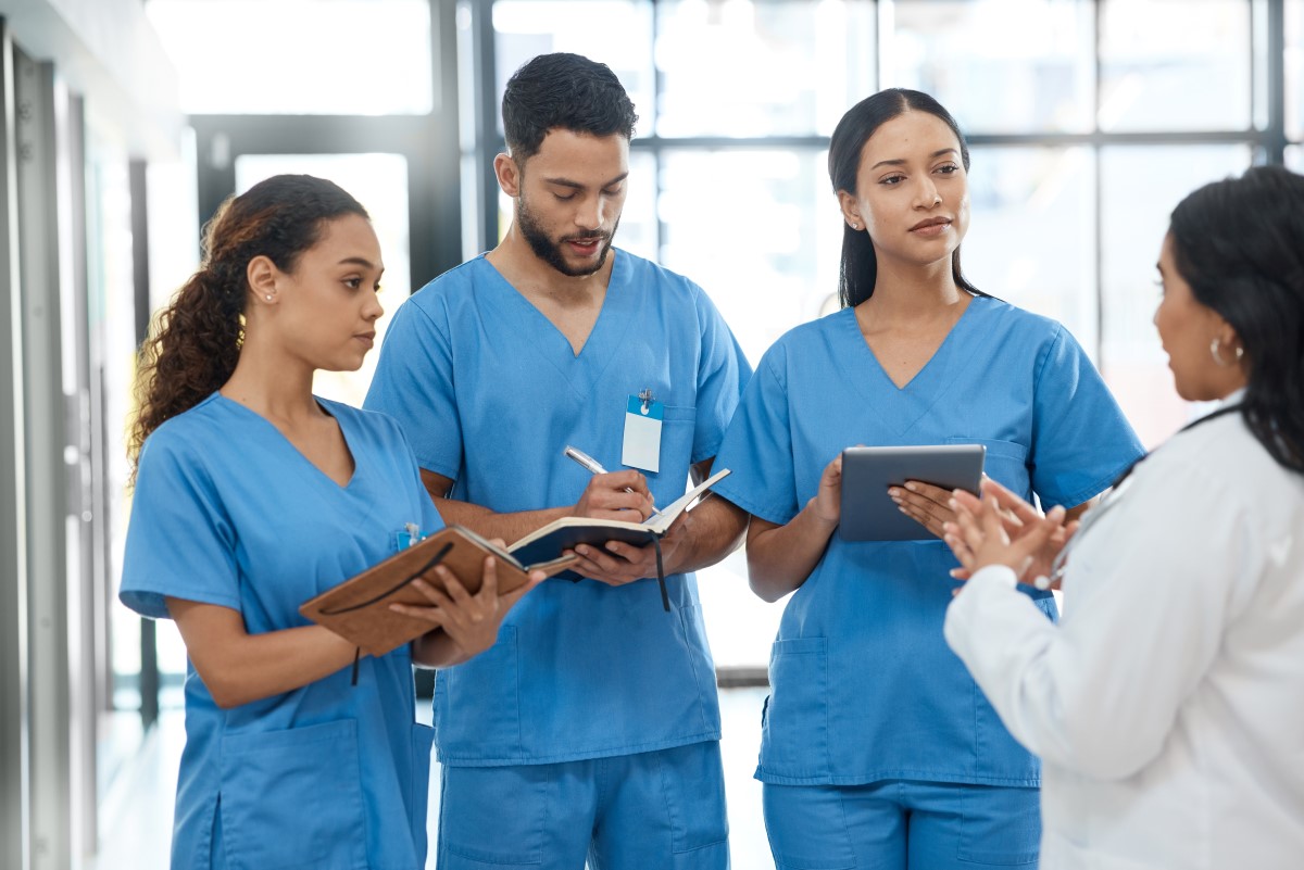 Three healthcare workers in blue scrubs take notes on clipboards and tablets while listening to a doctor in a white coat in a bright hospital hallway.