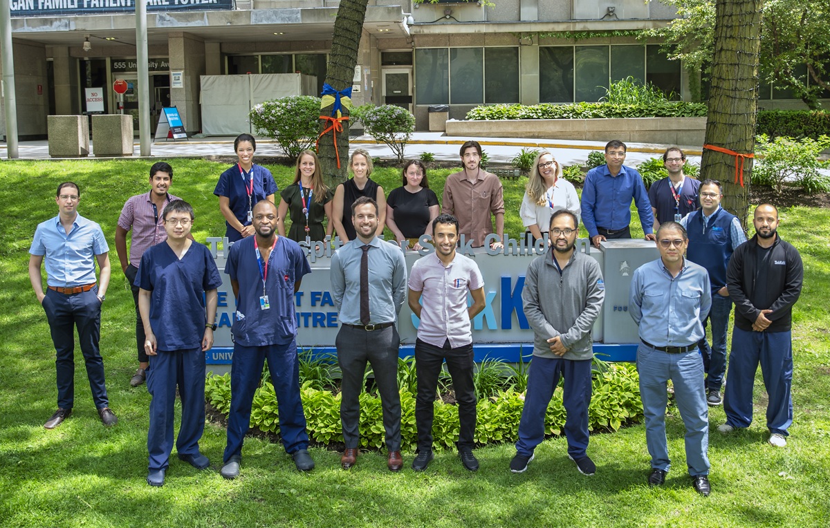 A group of fifteen people standing outdoors on grass in front of a hospital sign, wearing business casual and medical attire.