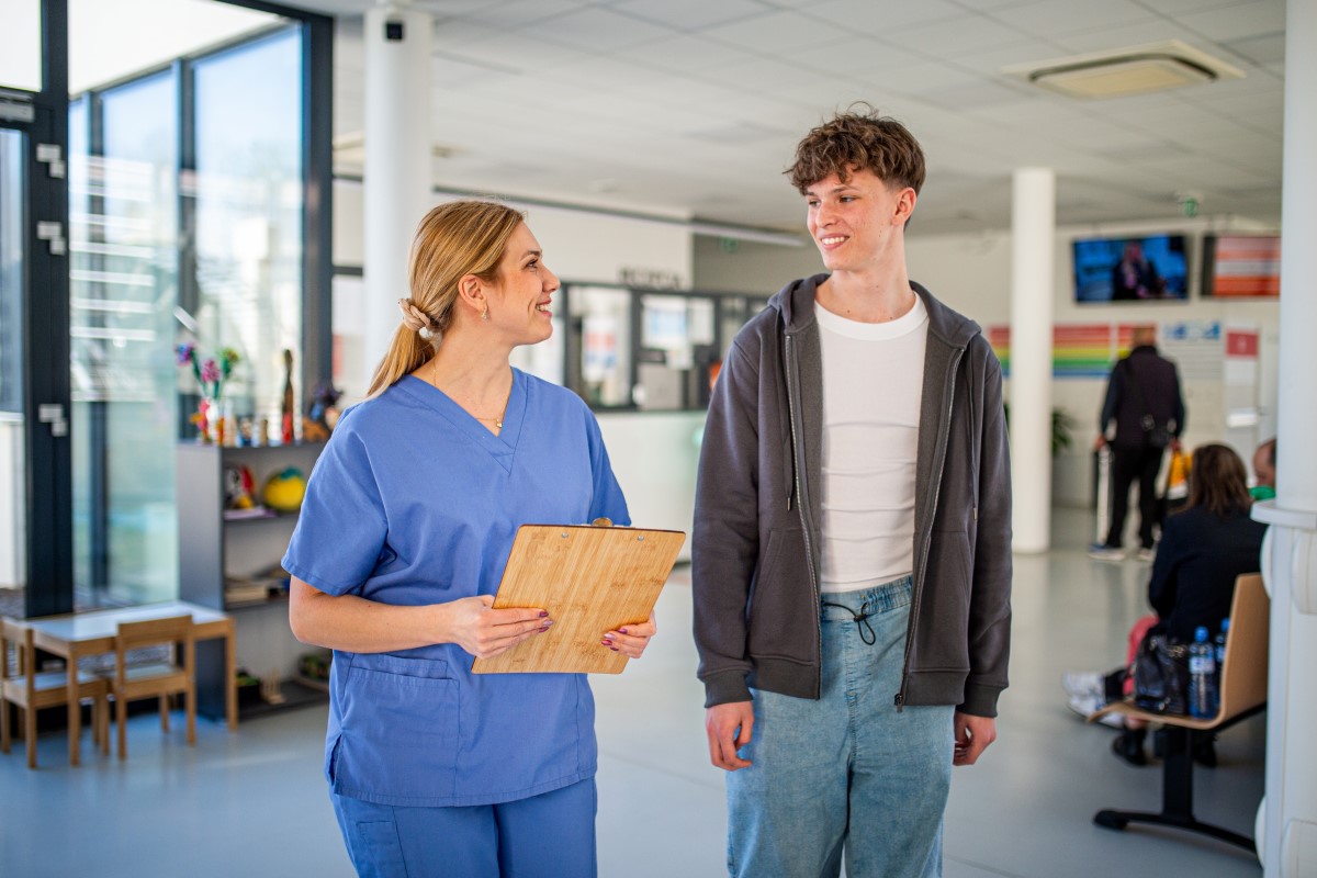 A healthcare worker in blue scrubs walks beside a young patient, holding a clipboard, inside a bright hospital waiting area.