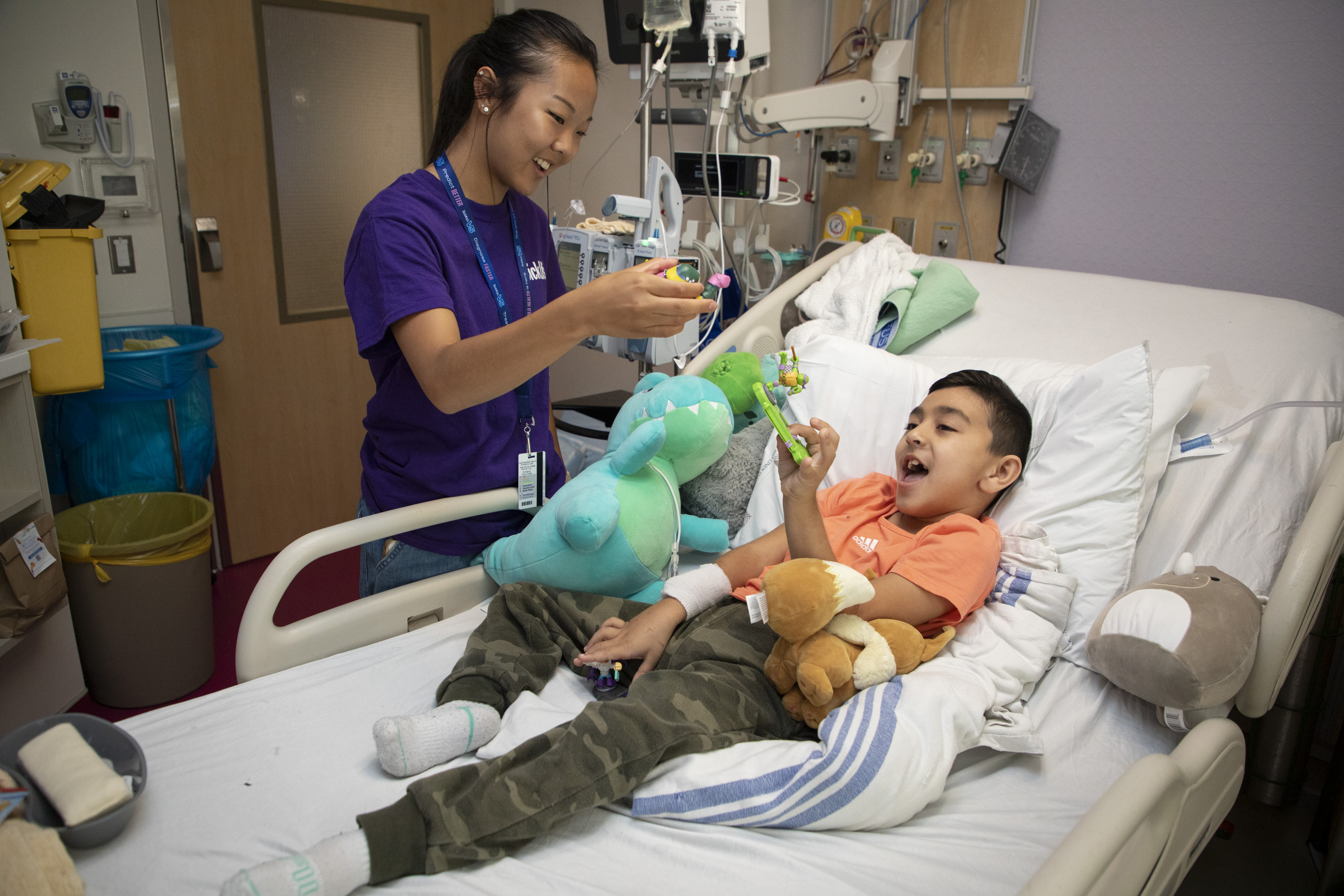 Teenager holds a toy, playing with a child in a hospital bed.