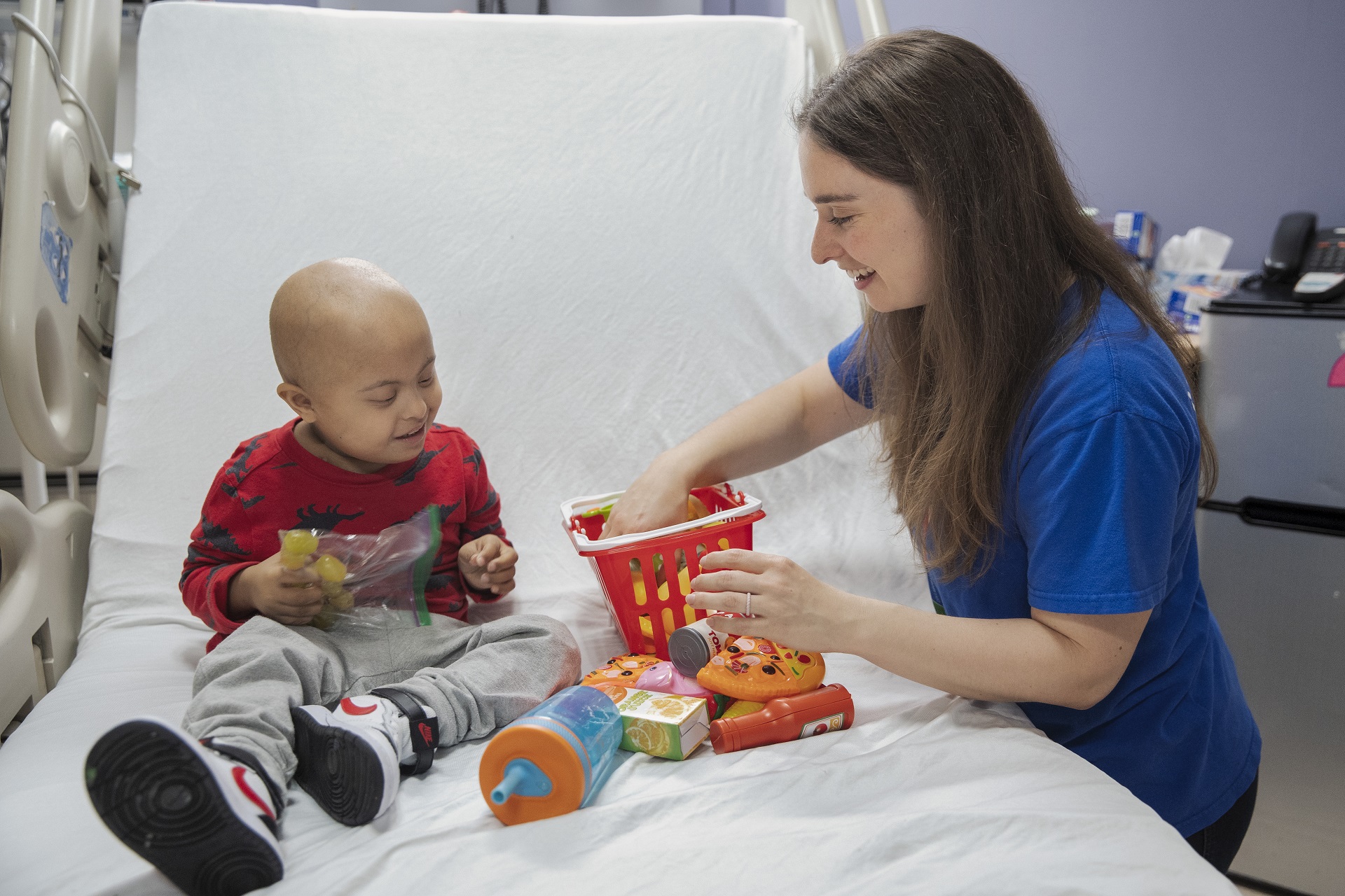 Child seated in a hospital bed plays with toy food, accompanied by a woman