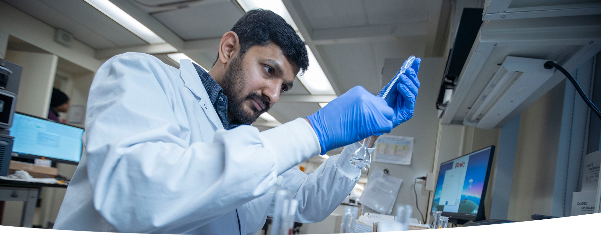 A SickKids staff member wearing a lab coat and blue gloves carefully pours a liquid from a pipette into a flask in a laboratory setting. He is focused on his task, with lab equipment and a computer screen visible in the background.