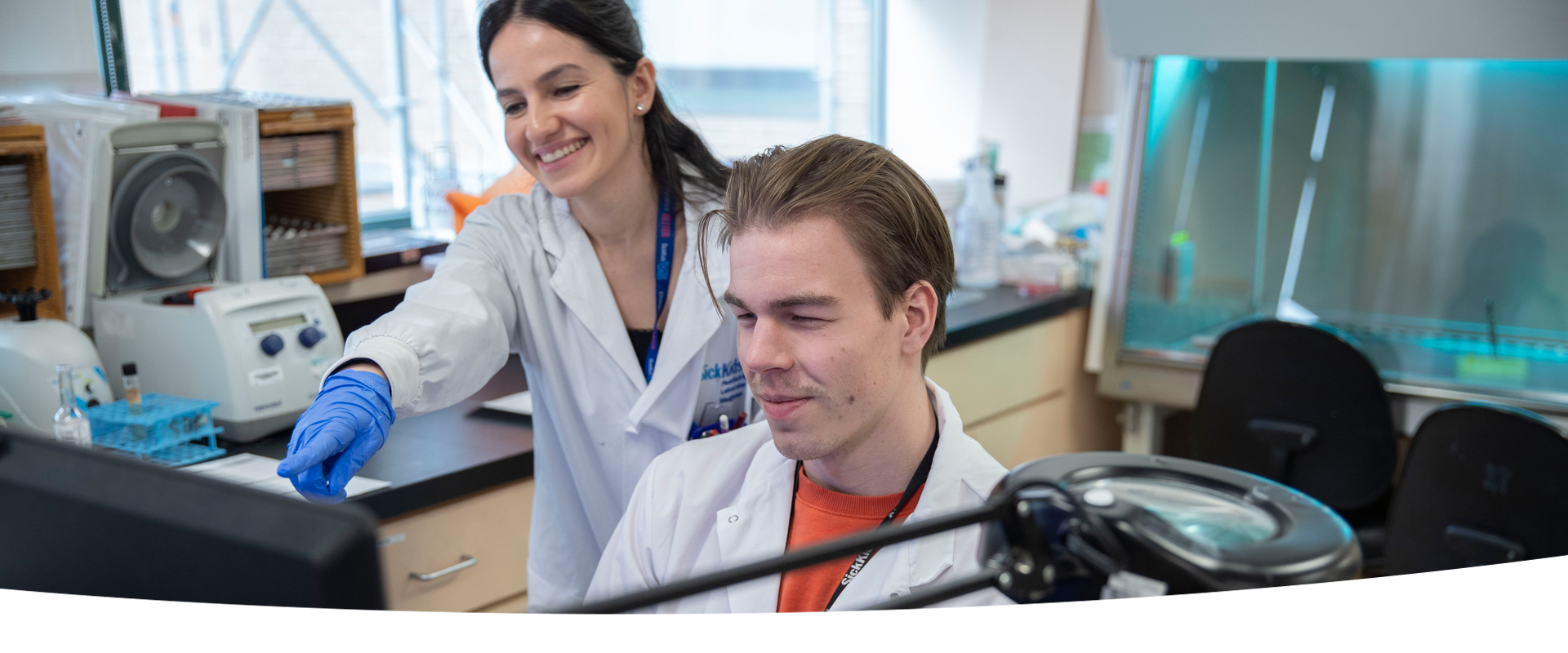 Two lab professionals at a lab bench. One is seated in front of a computer screen while the other is standing over and pointing at the screen.