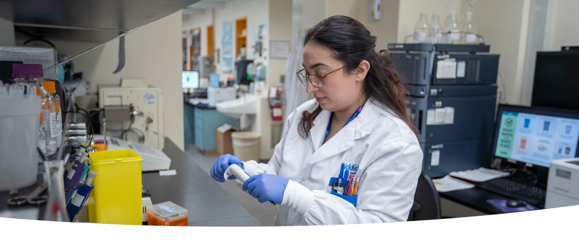 A lab professional wearing a lab coat and blue gloves. She is handling a piece of equipment at a lab bench.