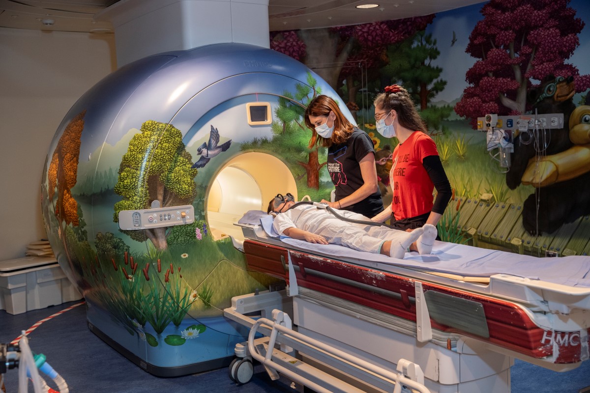 Two staff preparing a child for an MRI. The MRI machine and walls of the room are decorated with nature scenery.