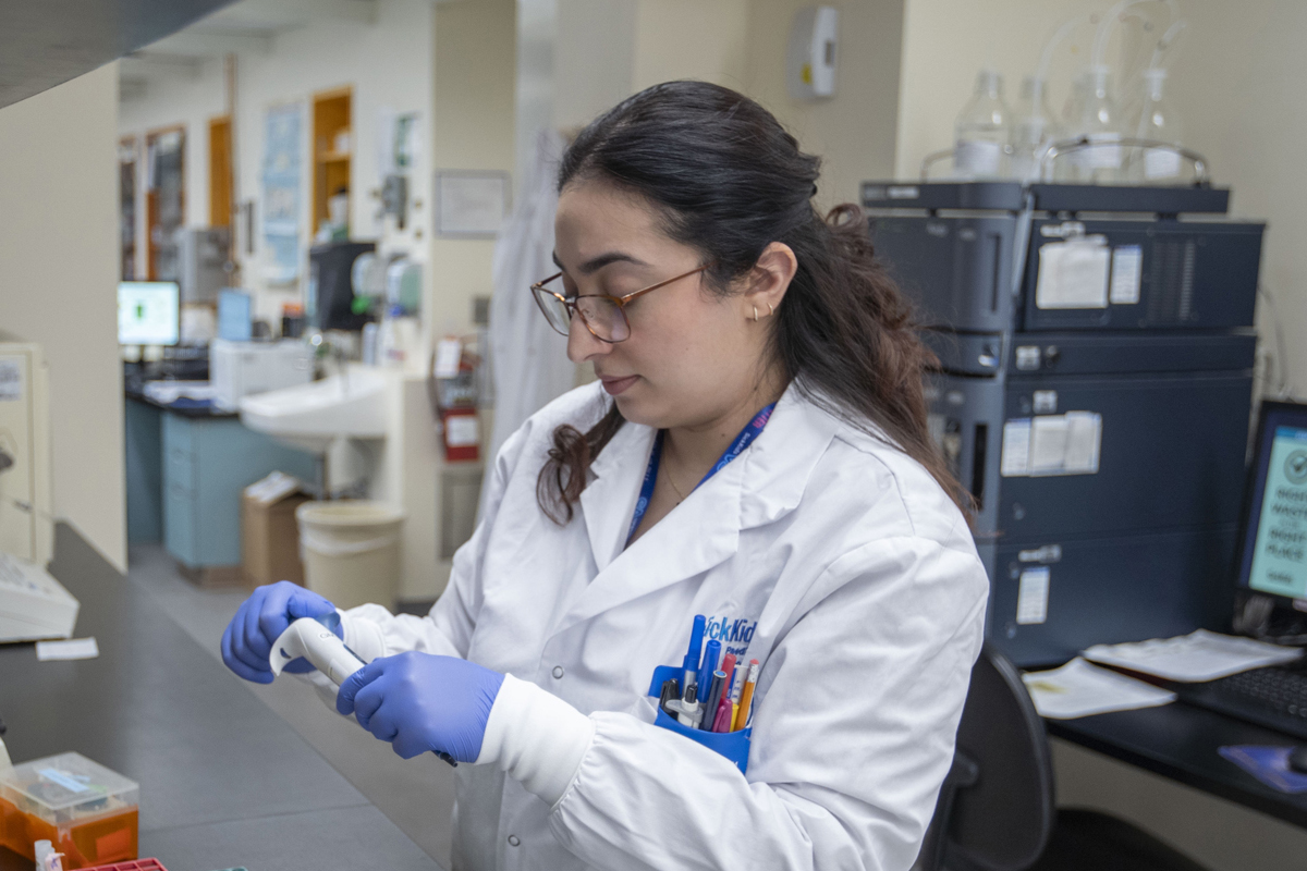 A SickKids staff member wearing a lab coat and blue gloves in a laboratory setting. She is handling a piece of equipment.