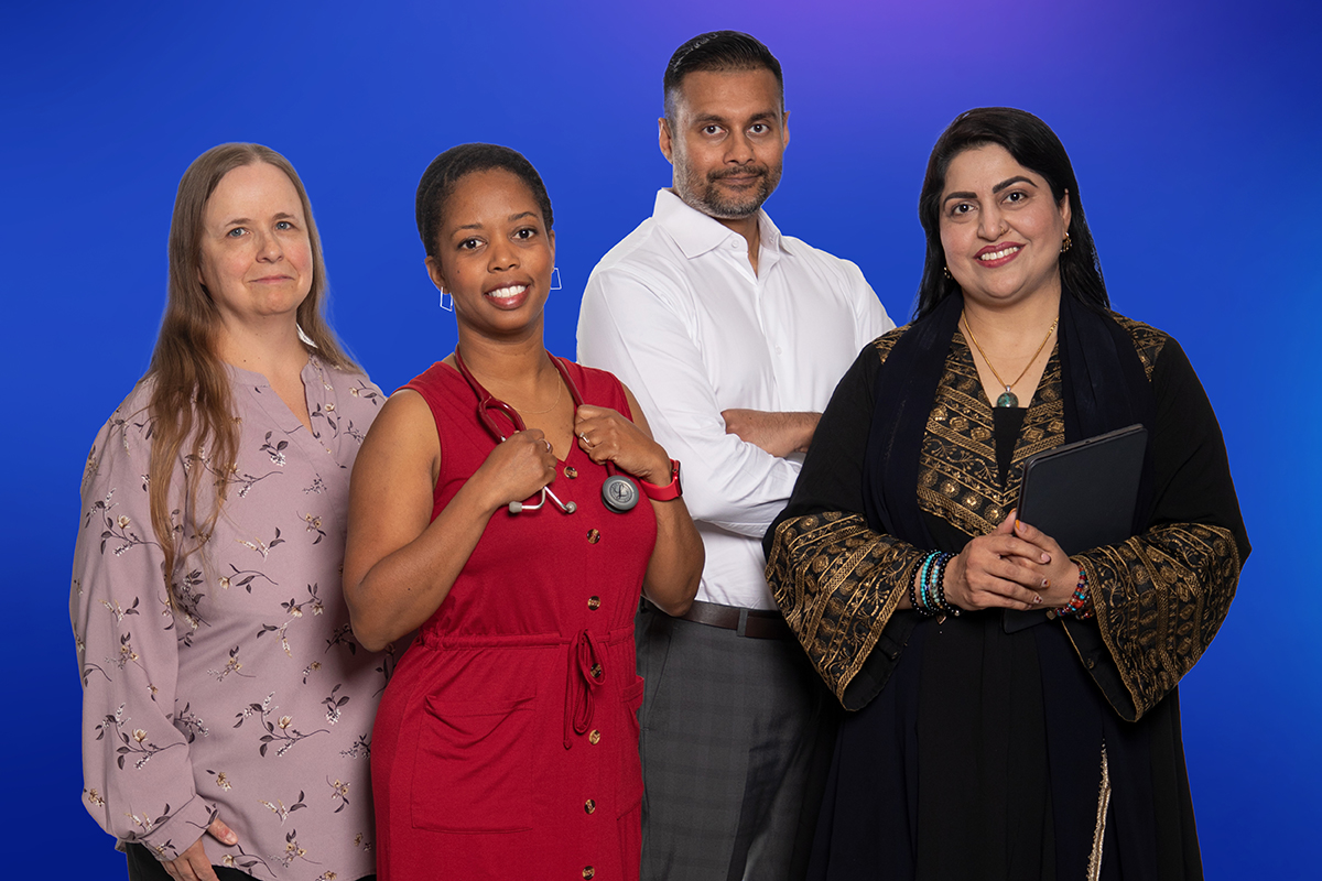 Four people standing together against a blue background. One person wears a light floral top, another wears a red dress with a stethoscope, a third wears a white shirt, and the fourth wears a black and gold patterned outfit holding a tablet.