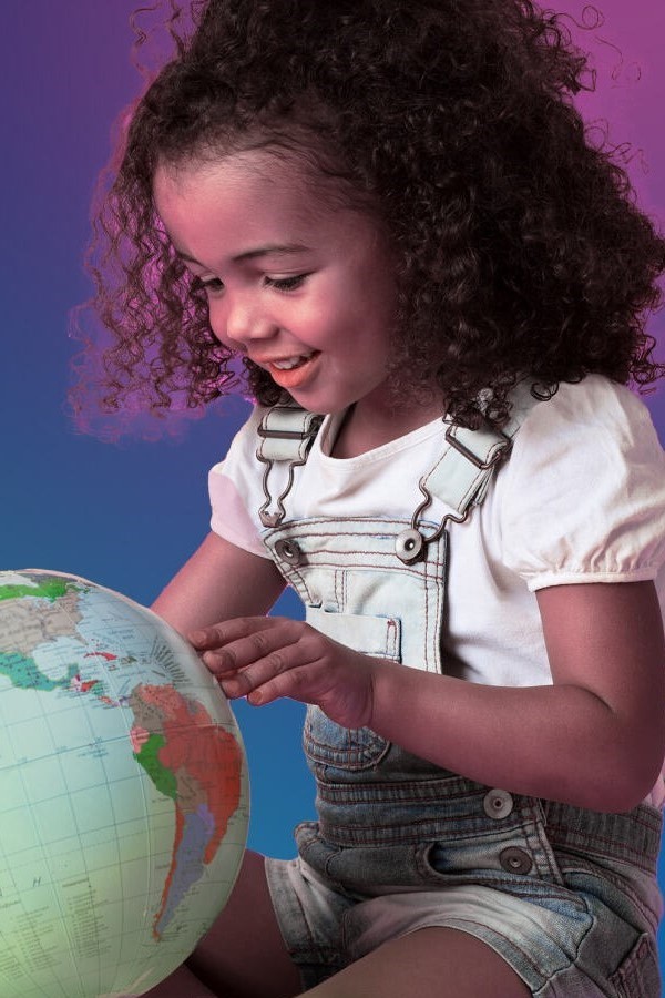 Young girl with curly hair in denim overalls smiling while exploring a colorful world globe against purple background.