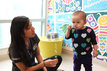 A woman wearing a SickKids Child Life tshirt and lanyard blows bubbles with a small child
