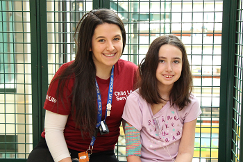 A woman wearing a SickKids Child Life tshirt and lanyard sites on a bench next to a young girl with an IV